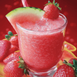 A refreshing watermelon strawberry smoothie in a clear glass, garnished with a slice of watermelon and a fresh strawberry, surrounded by ripe strawberries on a red background.