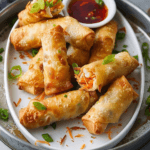 A plate of golden-brown crispy shrimp egg rolls, garnished with sliced green onions and served with a side of sweet and spicy dipping sauce. The egg rolls are filled with shrimp, shredded carrots, and cabbage.