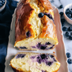 A freshly baked blueberry cream cheese bread with a golden crust, swirled with creamy cheese and juicy blueberries, served on a white platter.