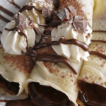 h Creamy chocolate-filled crepes topped with whipped cream, chocolate drizzle, and chocolate chunks on a white plate.