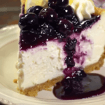 A slice of lemon blueberry cheesecake with a creamy filling, golden graham cracker crust, and a luscious blueberry topping, garnished with whipped cream and lemon slices.