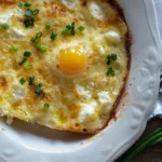 A golden, cheesy dish of baked cottage cheese eggs, topped with green onions and a perfectly baked egg in a white plate.