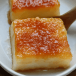 A close-up of two slices of creamy no-bake cassava cake with a caramelized golden top, served on a white plate with a wooden spoon.