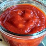 Close-up of a glass jar filled with thick, rich, homemade ketchup, showcasing its deep red color and smooth texture.