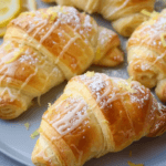 Golden, flaky lemon cheesecake crescent rolls drizzled with icing, dusted with powdered sugar, and garnished with fresh lemon zest on a plate.