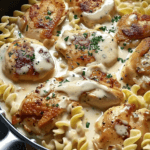 A skillet filled with golden-seared chicken in a rich, creamy sauce, served over tender buttered egg noodles and garnished with fresh herbs.