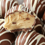 Close-up of no-bake peanut butter cheesecake balls coated in milk chocolate with white chocolate drizzle, revealing a creamy peanut butter filling.