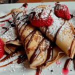 Golden crepes filled with Nutella, drizzled with chocolate sauce, topped with fresh strawberries, and dusted with powdered sugar.