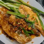 Pan-seared fish fillets topped with crispy garlic, fresh scallions, and a savory ginger-soy sauce, served on a white plate.