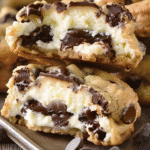 Close-up of a cheesecake-stuffed chocolate chip cookie cut in half, revealing a creamy cheesecake filling surrounded by gooey melted chocolate and golden-brown cookie dough.