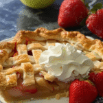 Slice of lattice-topped fruit pie with visible apple and rhubarb filling, topped with whipped cream and served with fresh strawberries.